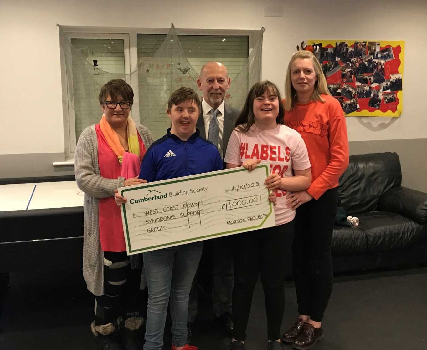 Five people stand indoors holding a large check for £1,000 made out to "West Coast Down's Syndrome Support Group" from Cumberland Building Society.