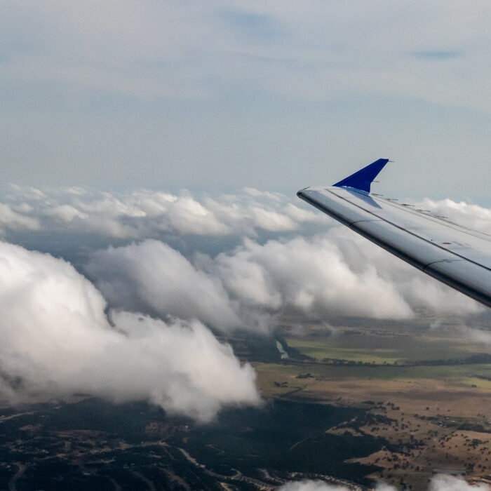 View from an airplane window shows a wing against a backdrop of scattered clouds, with fields and landscape visible below.