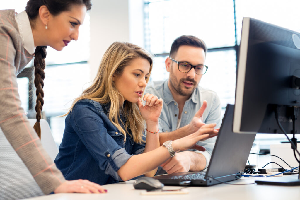 Three people work together at a desk, looking at a computer monitor and discussing something on the screen in an office setting.