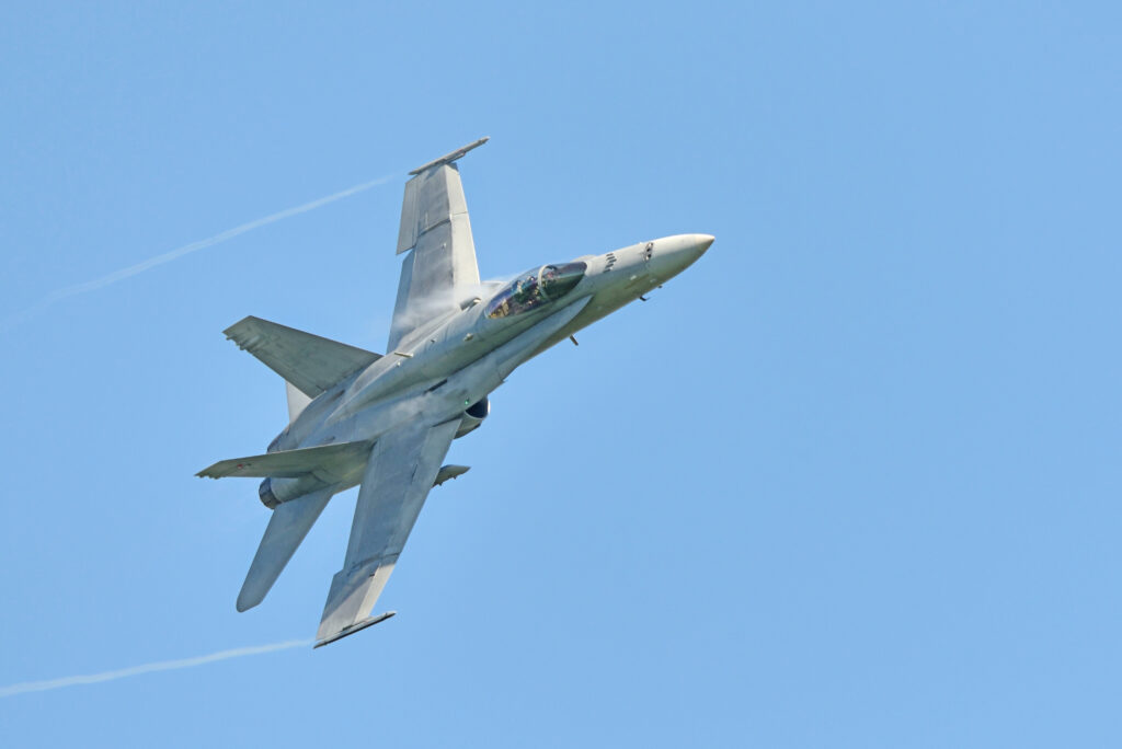 A gray fighter jet flies at an angle against a clear blue sky, leaving faint condensation trails behind its wings.