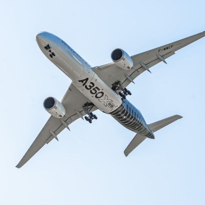 A commercial Airbus A350 XWB aircraft is flying overhead against a clear blue sky, with landing gear extended.
