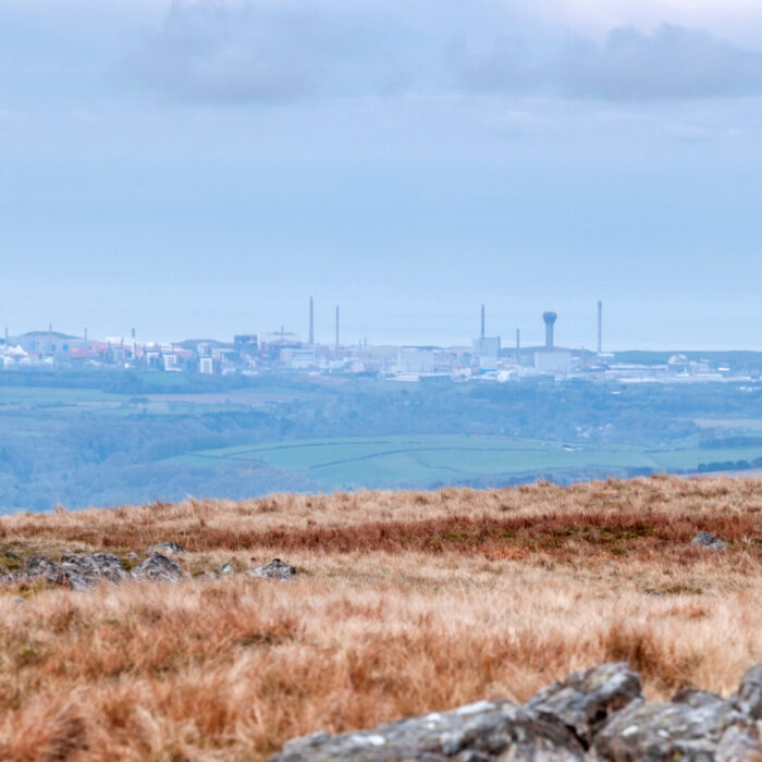 Rocky, grassy moorland in the foreground with an industrial facility and tall chimneys visible in the distance under a cloudy sky.