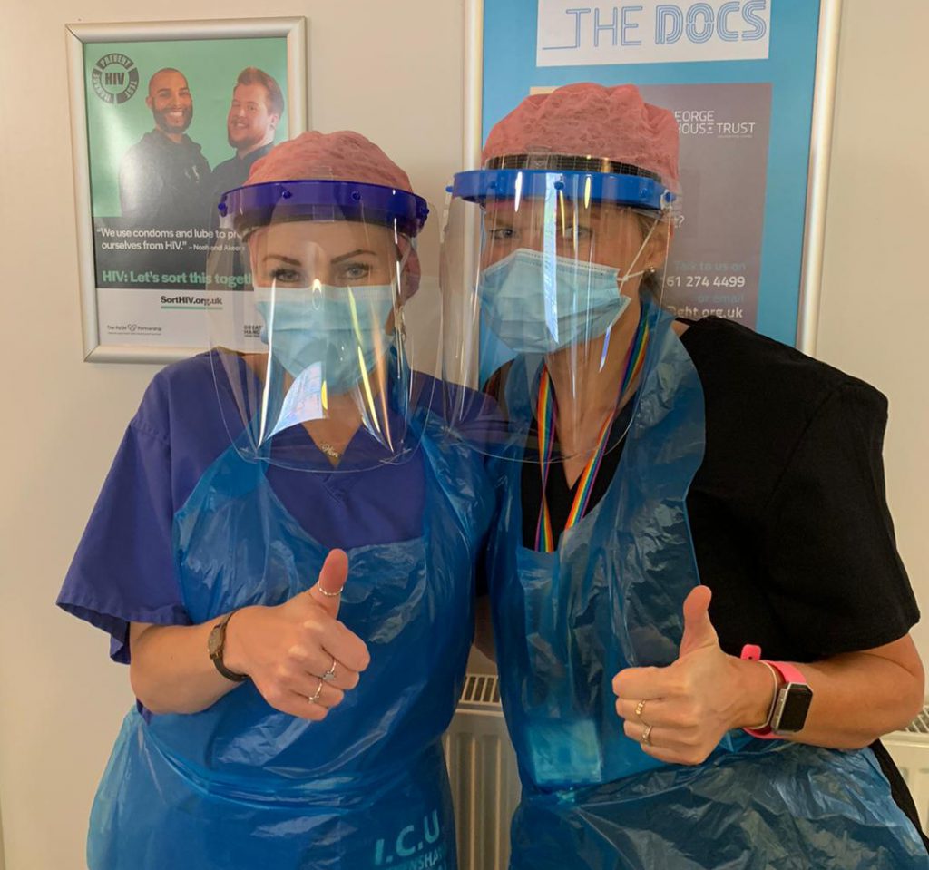 Two healthcare workers wearing masks, face shields, scrubs, and hair covers give a thumbs-up gesture while standing indoors.