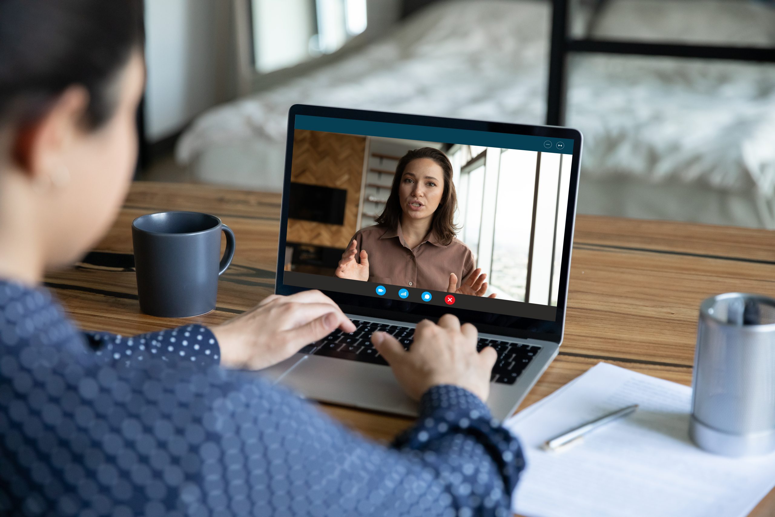 Person participating in a video call on a laptop, with a woman speaking on the screen; a mug, notebook, and pen are on the desk.