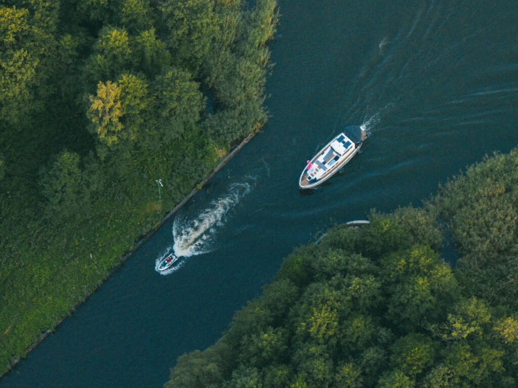 Aerial view of a narrow river bordered by dense trees, with a small motorboat and a larger yacht traveling in opposite directions.