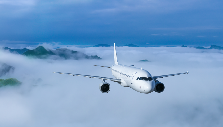 A white commercial airplane flies above clouds with mountain peaks visible in the background under a blue sky.