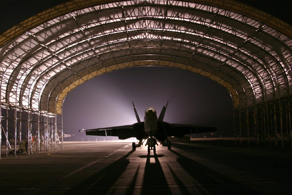 A fighter jet is parked under a large arched hangar at night, silhouetted by bright lights shining from behind.