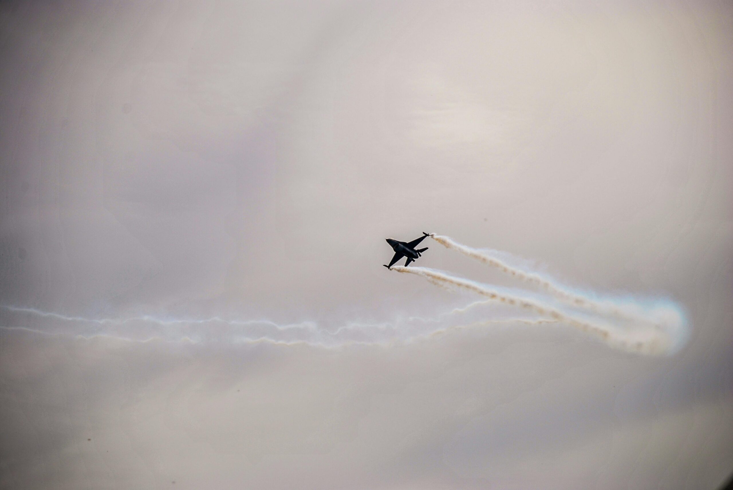 A jet fighter flies through a cloudy sky, leaving visible vapor trails behind its wings.