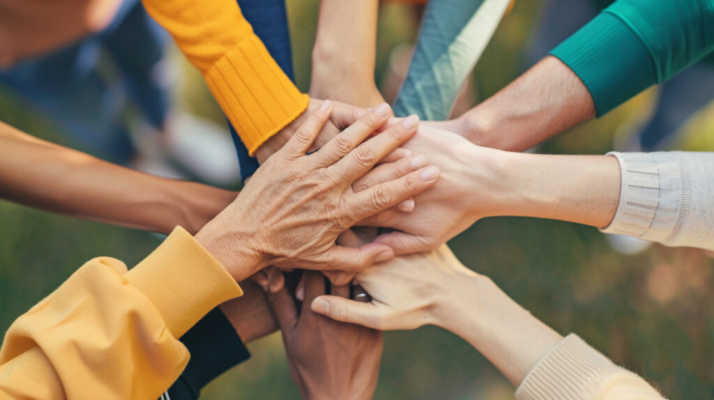 A group of diverse hands stacked together in a circle, symbolizing teamwork and unity, with various skin tones and clothing visible.
