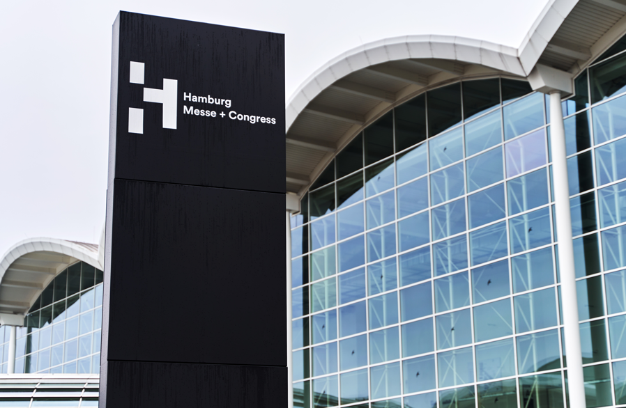 A black sign reading "Hamburg Messe + Congress" stands in front of a modern glass building with a curved white roof.