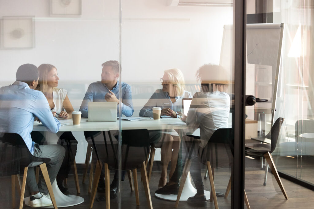 Five people sit around a conference table in an office, engaged in discussion with laptops, papers, and coffee cups visible.