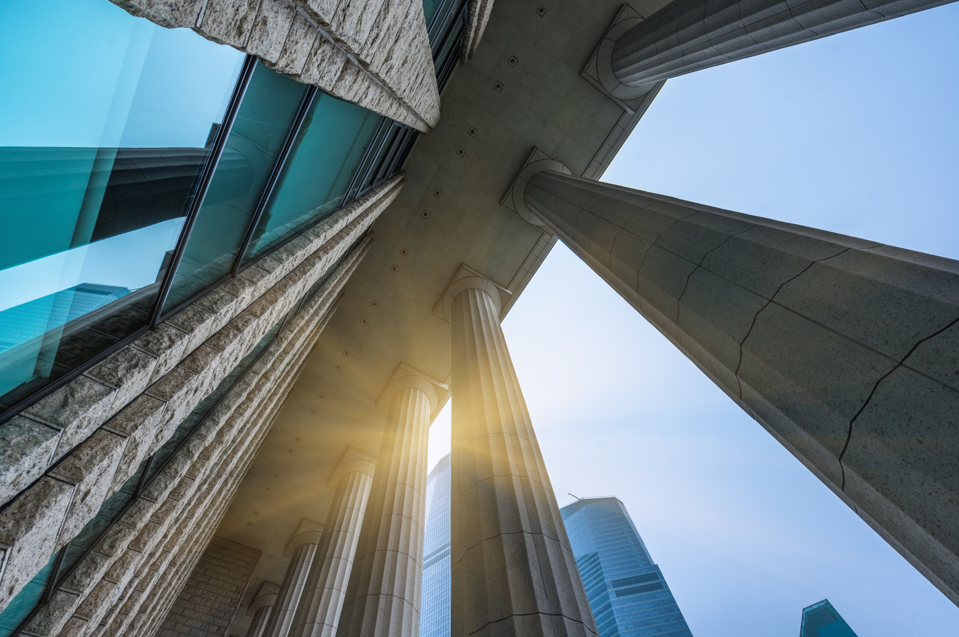 Low-angle view of tall stone columns and a modern glass building with sunlight shining through, against a blue sky and city skyscrapers in the background.