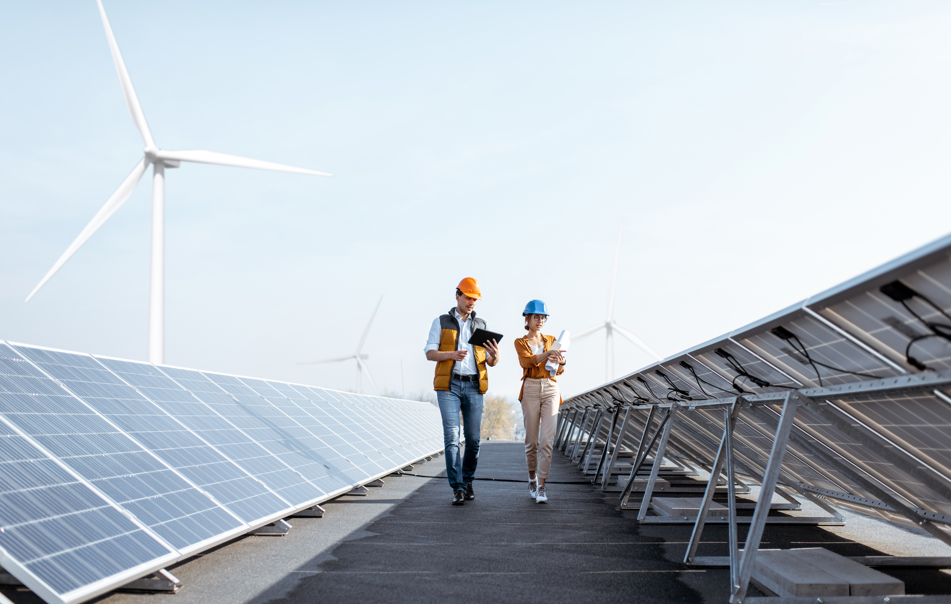 Two engineers wearing hard hats inspect solar panels on a rooftop with wind turbines in the background on a clear day.
