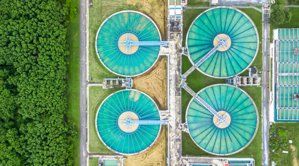 Aerial view of four large circular water treatment tanks next to a green forested area, separated by a road.