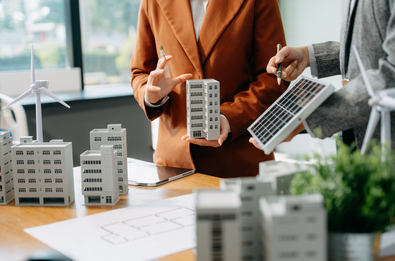 Two people discuss sustainable architecture using model buildings, a solar panel, and wind turbines on a table, with blueprints and greenery in the workspace.