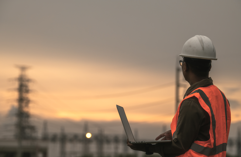 A person wearing a hard hat and a reflective vest works on a laptop outdoors near electrical infrastructure at sunset.