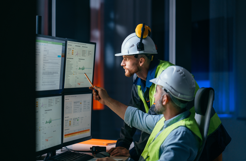 Two engineers wearing hard hats and safety vests view data on multiple computer monitors in an industrial control room; one points at the screen.