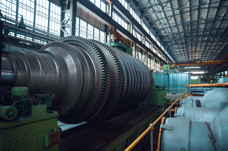 Large industrial turbine with gears situated inside a spacious factory, surrounded by machinery and metal structures under a high ceiling with windows.