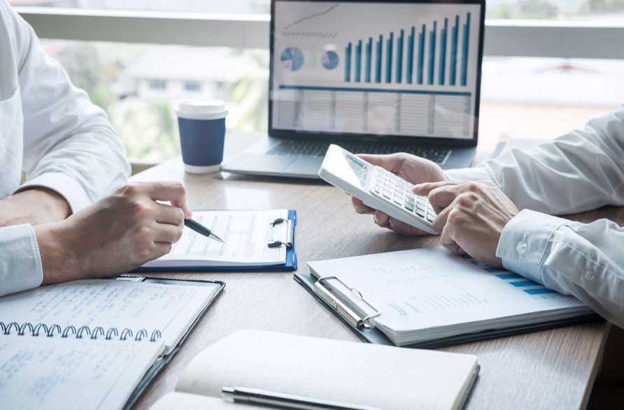 Two people working at a desk with documents, a calculator, notebooks, and a laptop displaying financial charts and graphs.