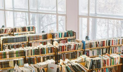 Rows of bookshelves filled with books in a library, with large windows in the background letting in natural light.