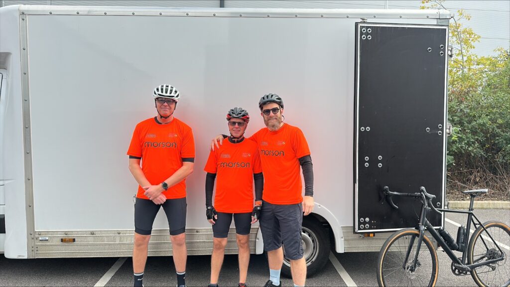 Three cyclists wearing matching orange jerseys stand in front of a white box truck, with a black bicycle visible on the right side.